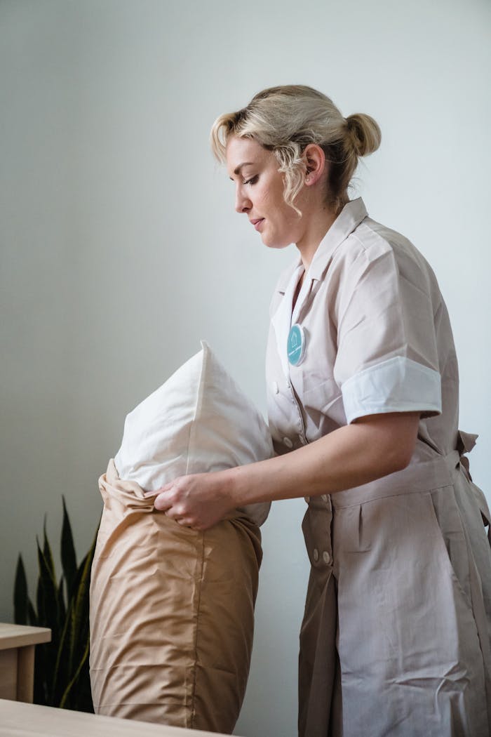 Hotel maid in uniform arranging pillows in a neatly styled guest room.